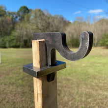 Load image into Gallery viewer, A 3/8in AR500 1x2 hook for mounting on 1x2 wood, placed outdoors with grassy background intended for steel shooting targets.